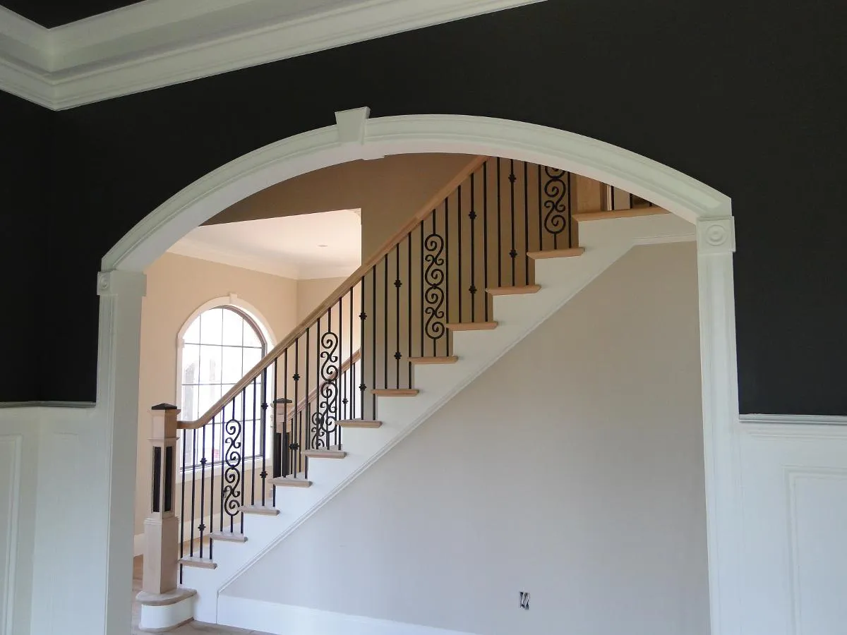 Staircase with iron balusters viewed through an arched radius-trim opening against a dark wall.
